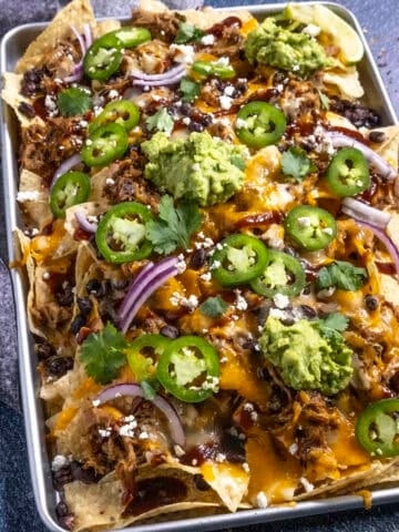 Serving up pulled pork nachos on a large tray, loaded with toppings, including guacamole, sliced jalapenos, sliced red onion, and bbq sauce